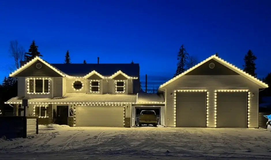 A house and detached garage outlined with warm white Christmas lights against a deep blue evening sky.