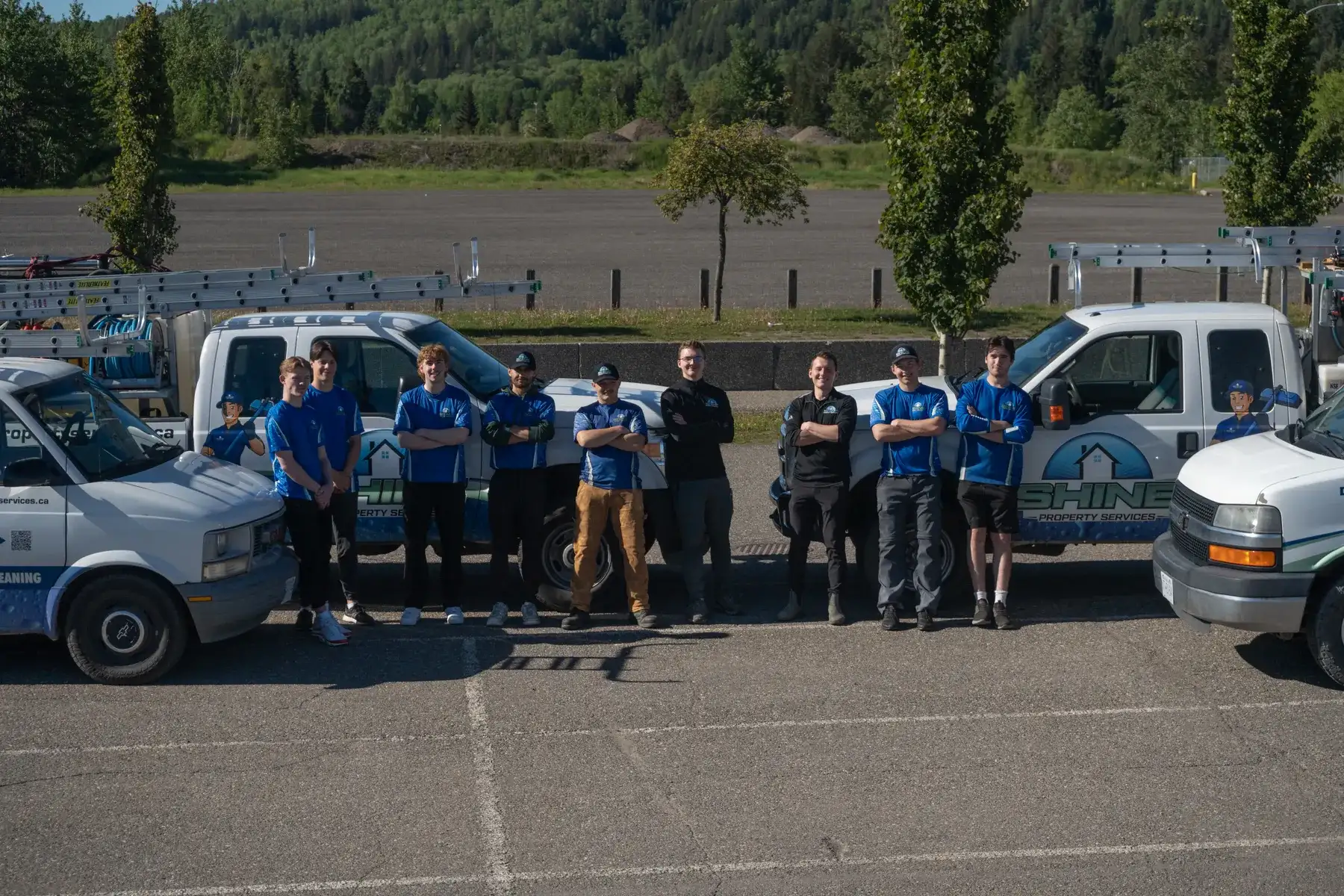 Eleven Shine Property Services crew members in blue branded shirts standing in a line in front of four branded work vans and pickups in a car park, forested hills behind.