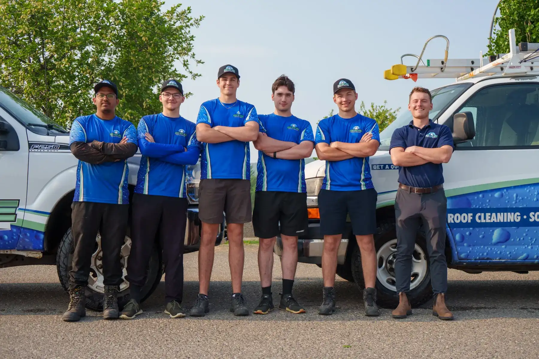 Six Shine Property Services crew members in blue branded shirts and caps standing with arms folded in front of two branded work vehicles with a ladder rack.