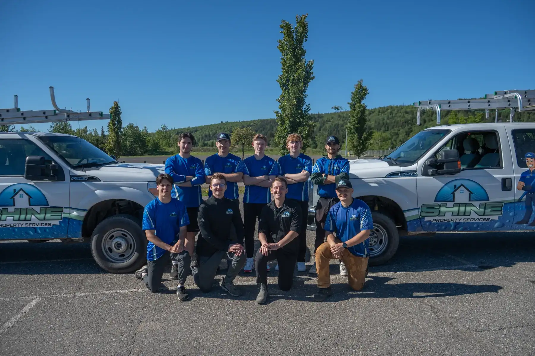 Nine Shine crew members in blue branded shirts, five standing and four kneeling, posed between two white branded F-250 pickups with ladder racks.