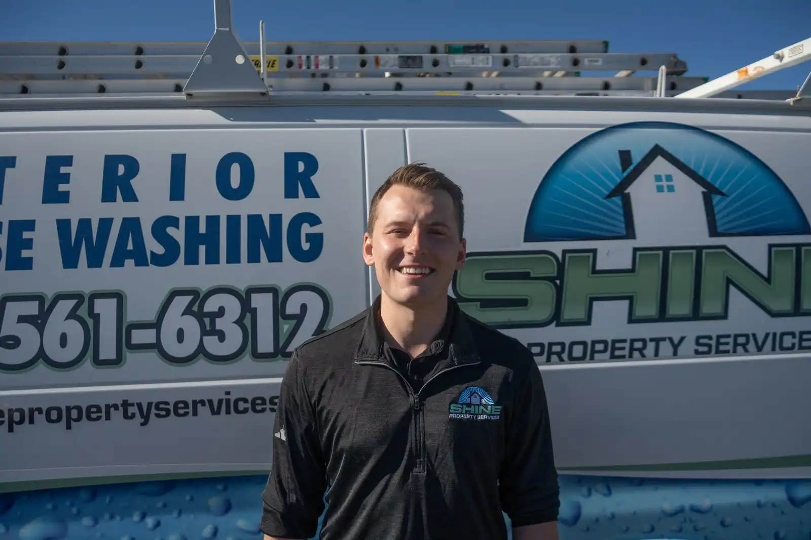 Craig MacDonald, founder of Shine Property Services, in a dark branded quarter-zip standing and smiling in front of a white work van lettered for exterior house washing.