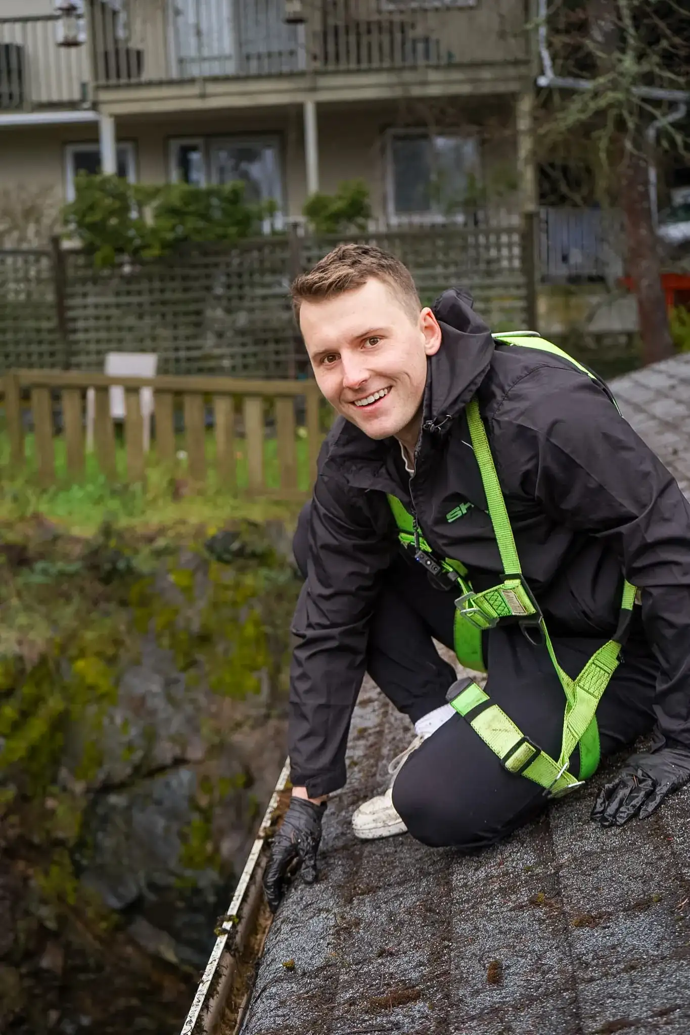 A Shine technician in a green harness leaning over a roof edge and clearing debris out of a gutter by hand.