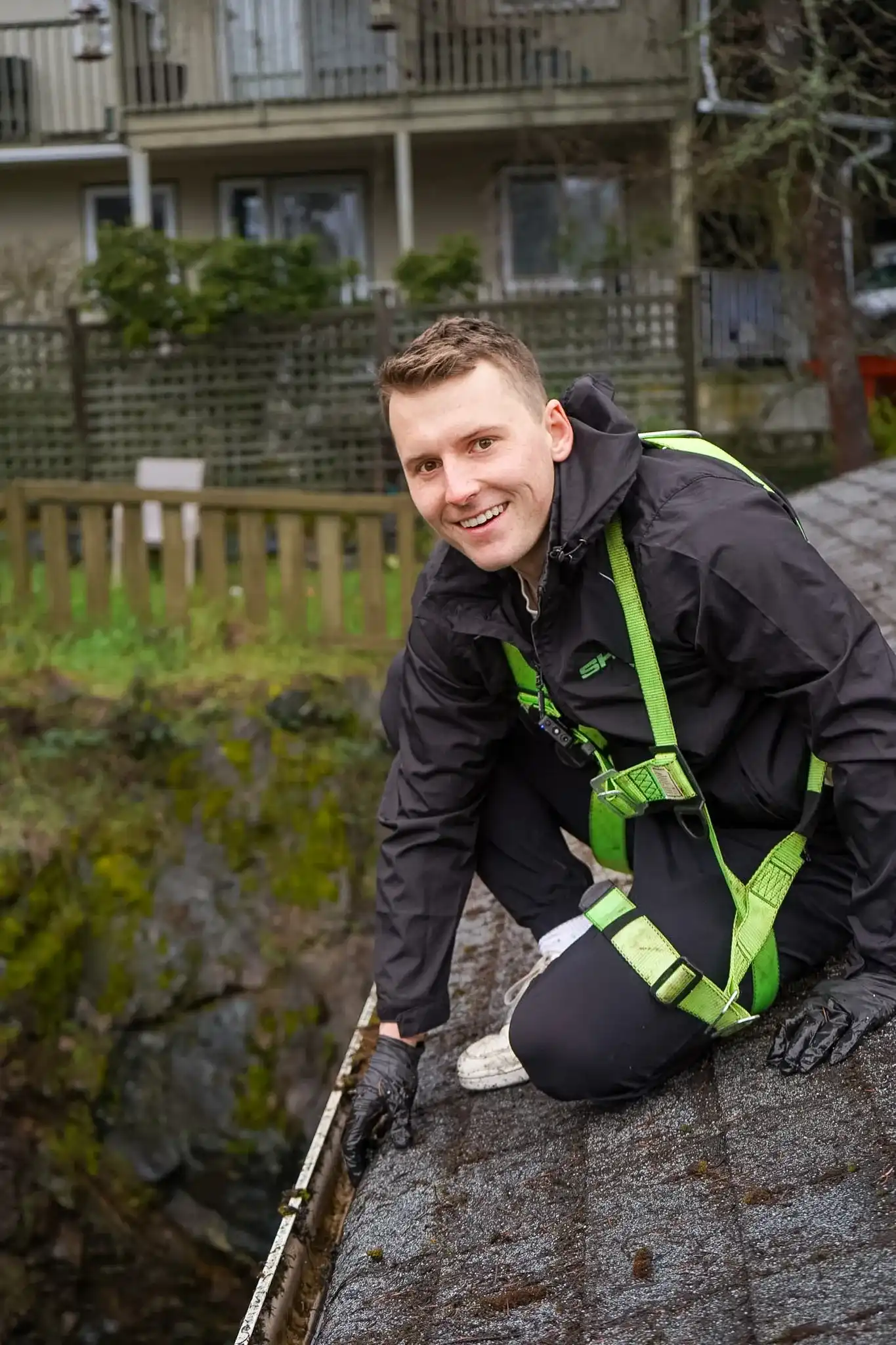 A Shine technician in a green harness leaning over a roof edge and clearing debris out of a gutter by hand.