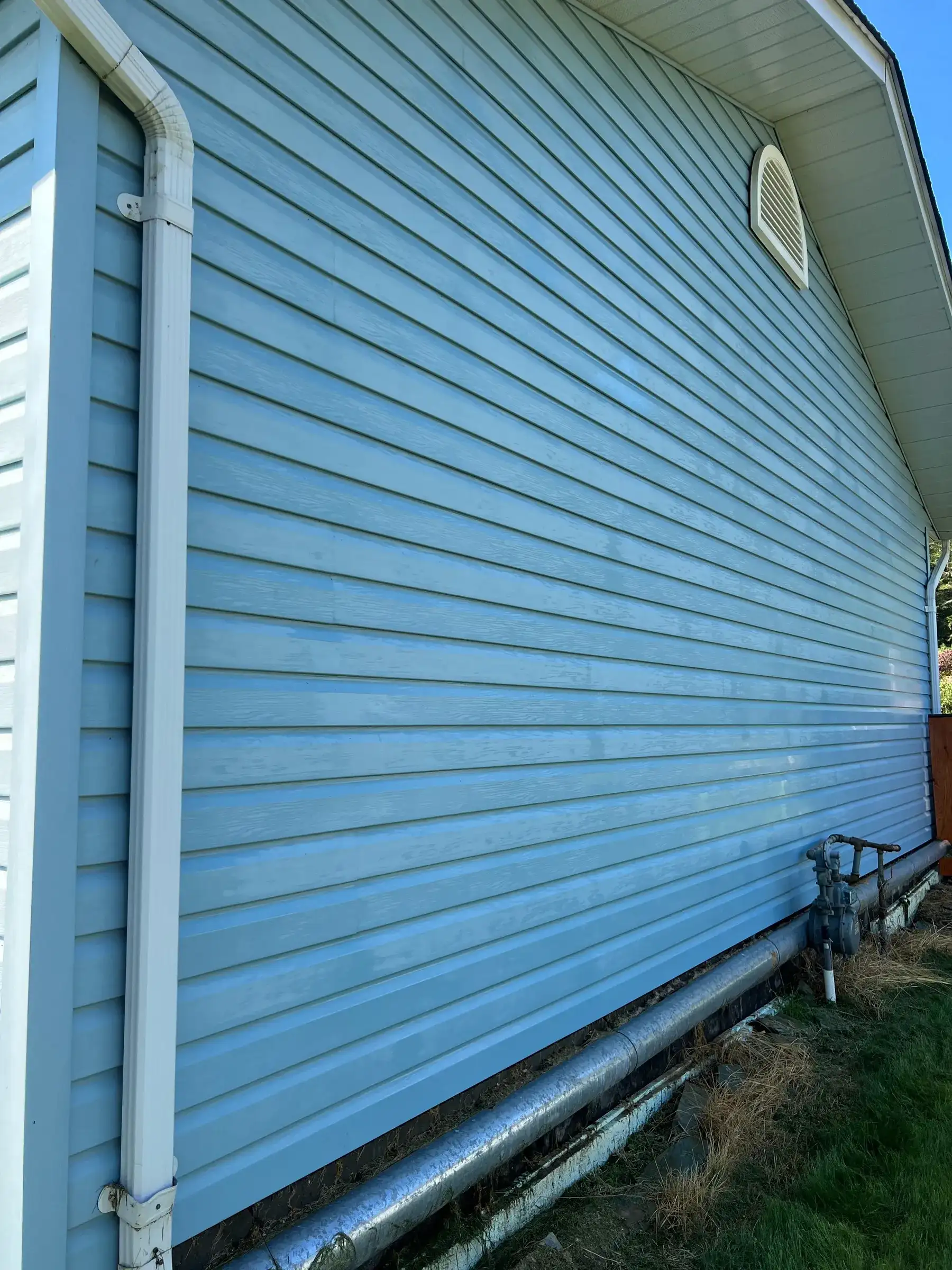 The same pale blue siding after soft washing, the streaking gone and the colour even, with the same downspout and gable vent in frame.