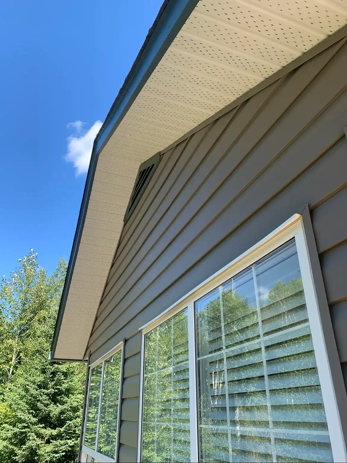 The same tan soffit and siding after soft washing, clean and even, with the same gable vent and window in frame under a clear blue sky.