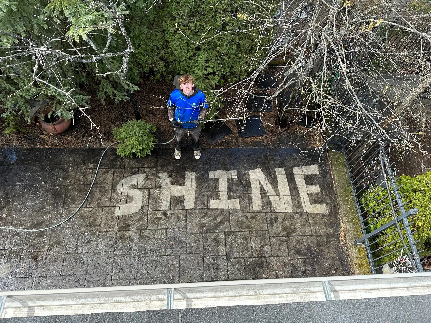 Looking down from a roof at a Shine technician with a pressure washer on a dirty stamped-paver patio, having cleaned the word SHINE into the surface in clean stone.