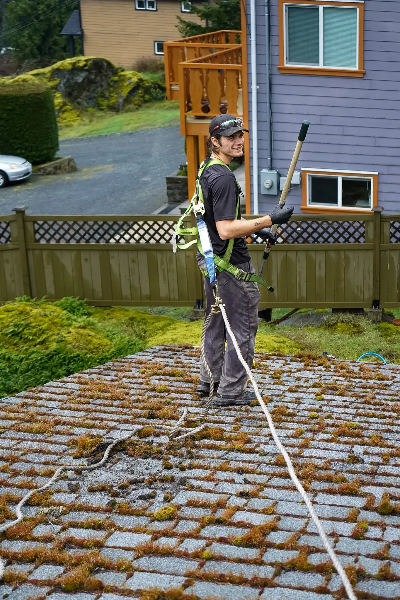A Shine technician standing in a harness and rope line on a roof thickly covered in moss, holding a long-handled brush and looking back over his shoulder.